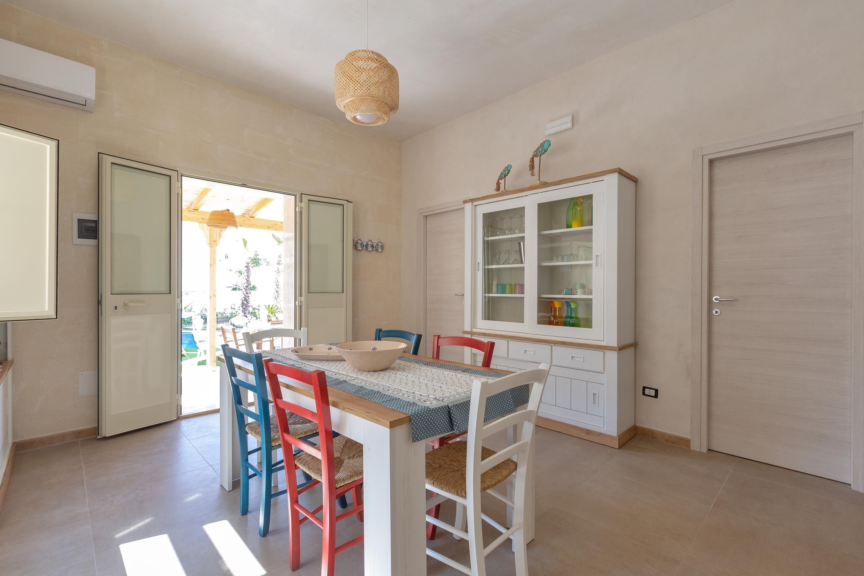 Un'area pranzo con tavolo apparecchiato per quattro persone, circondata da sedie colorate, un pensile con vetri e un lampadario.