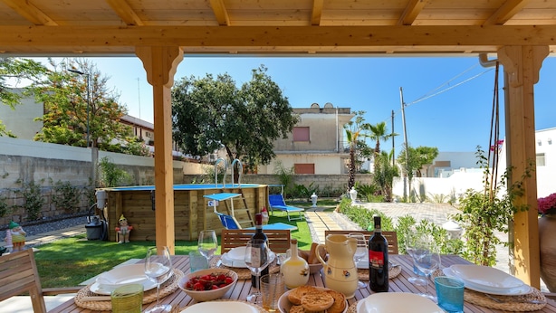 Un espace de restauration en plein air agrémenté d’une pergola en bois, avec des tables dressées : assiettes, verres et une bouteille de vin y sont disposés.