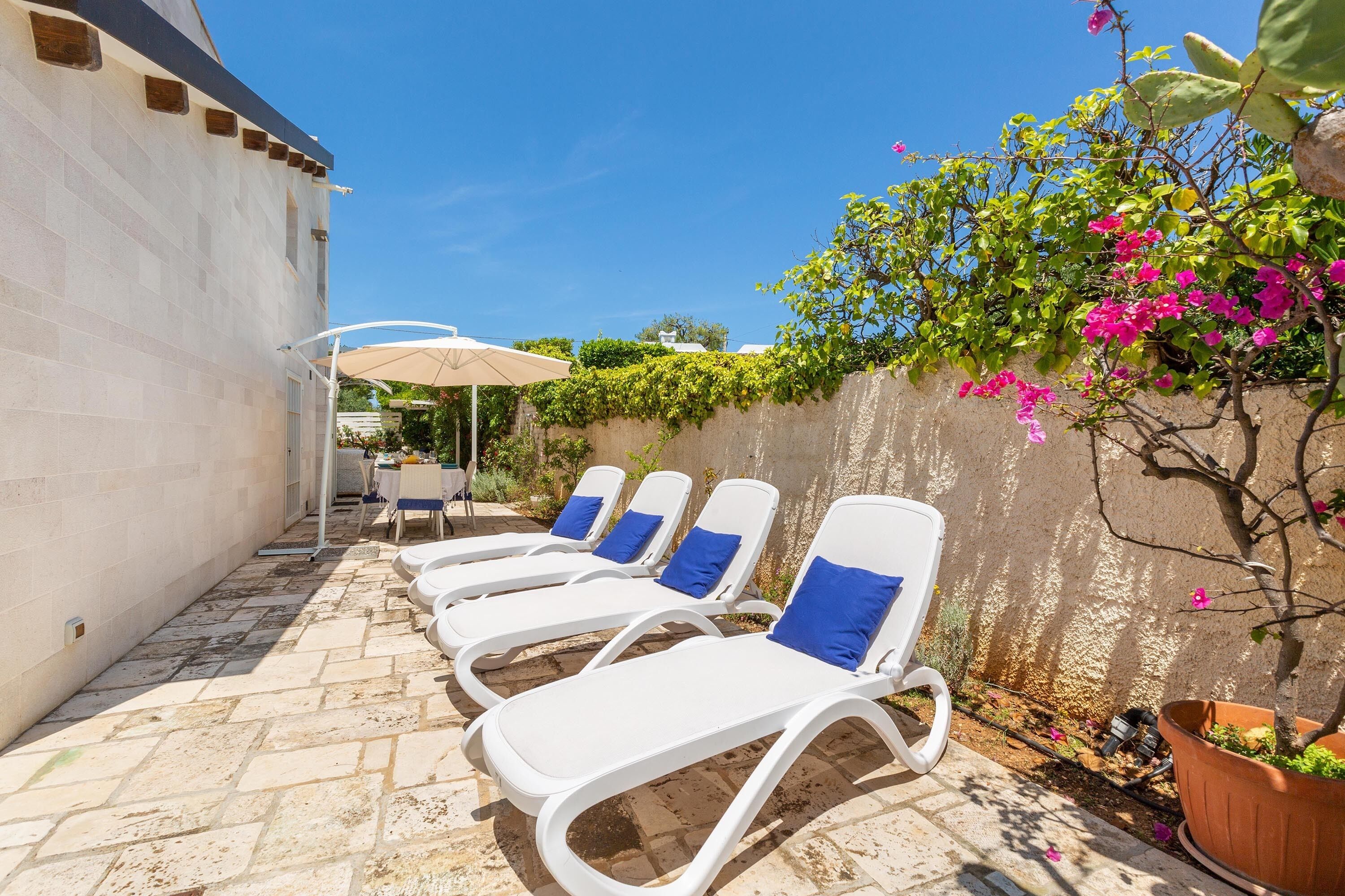 Une terrasse avec des fauteuils de jardin blancs et une table, entourée d’un mur de pierre et de plantes en pot.