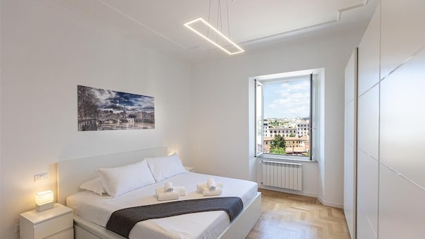 A modern bedroom with a bed, bedside tables, a window with a city view, and a painting on the wall.