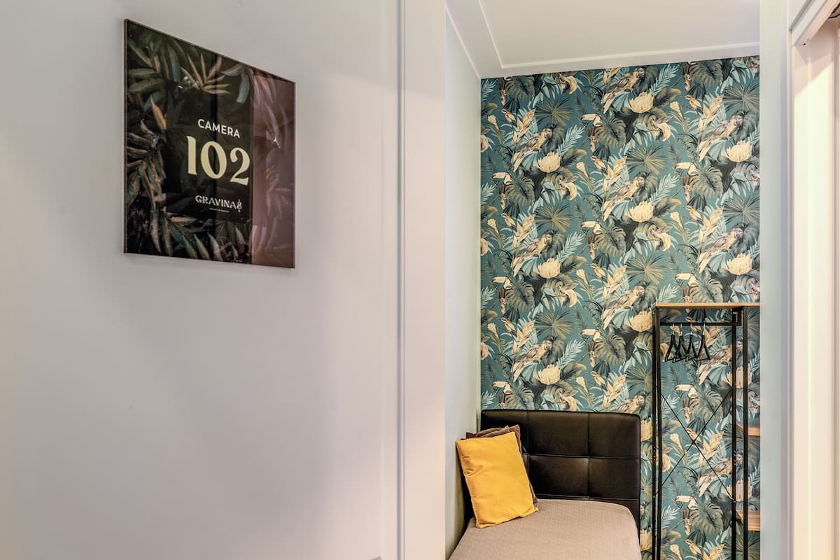 A bedroom with a bed, a headboard, a wall with a tropical leaf pattern, and a framed picture on the wall.