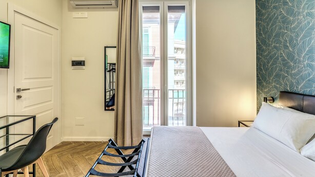 A hotel room with a bed, a desk, a chair, an air conditioning unit, a window with curtains, and a patterned wall. - Gravina8 (Naples)