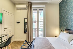Une chambre d’hôtel comprenant un lit, un bureau, une chaise, un appareil de climatisation, une fenêtre avec des rideaux et un mur décoré d’un motif.
