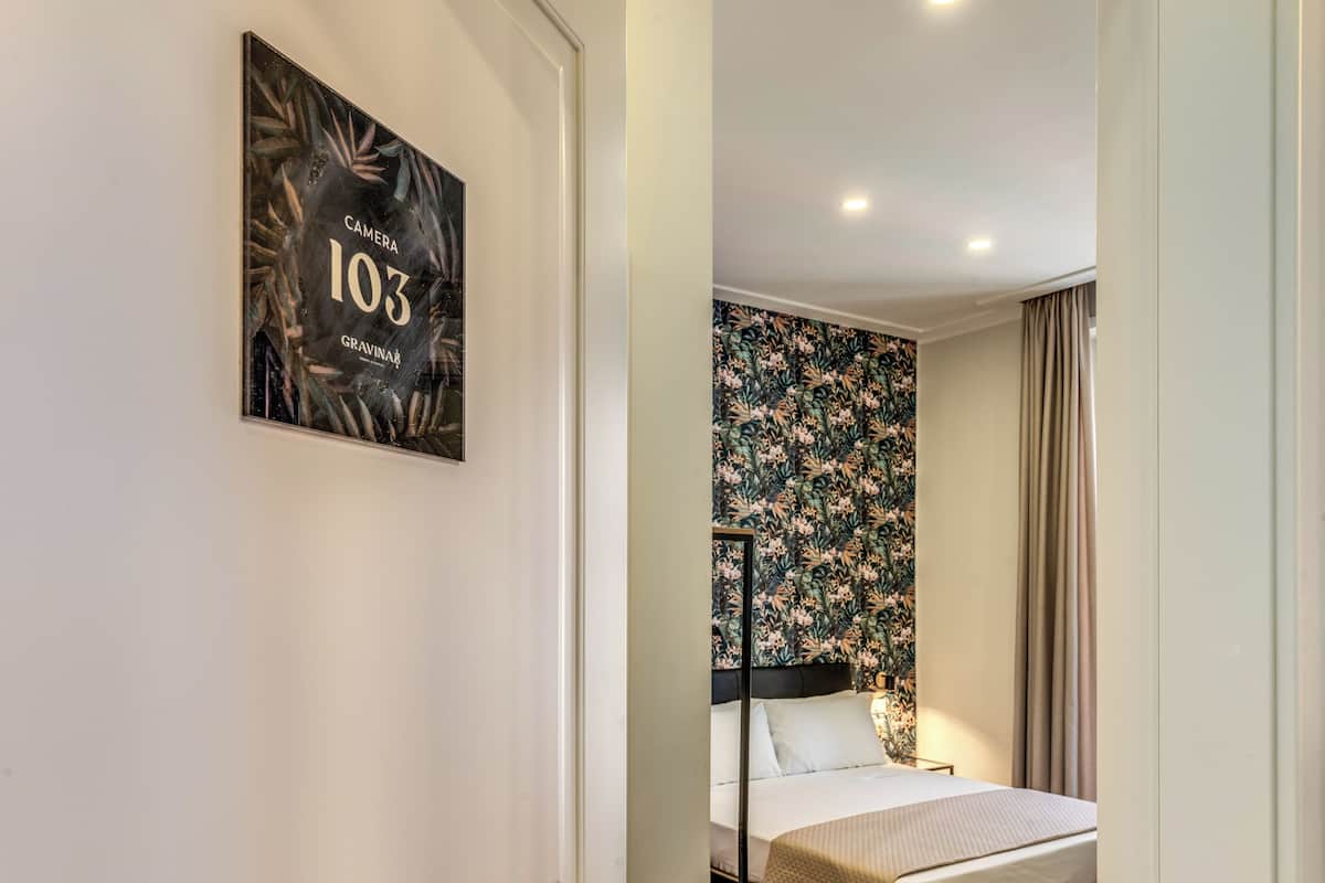 A hotel room with a bed, a headboard, a floral wallpaper, and a framed picture on the wall.