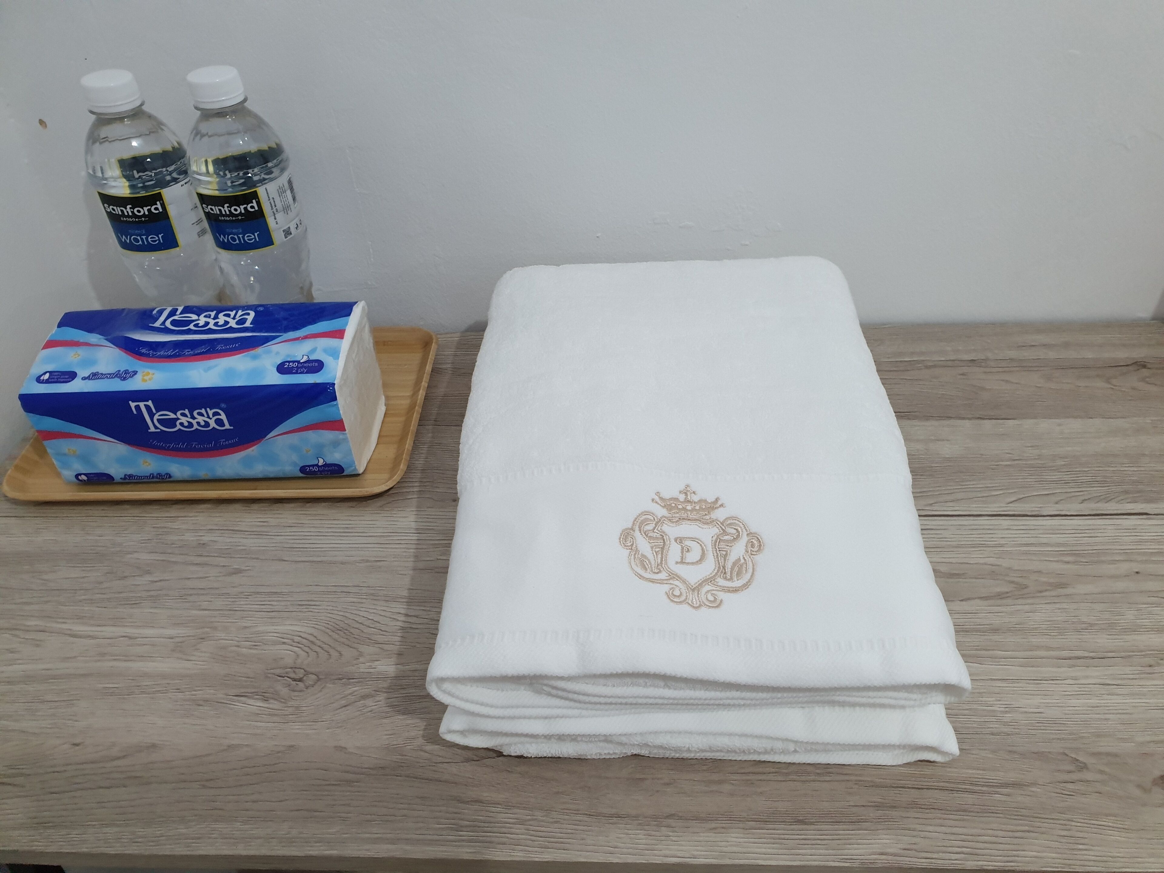 Una mesa de madera con una toalla blanca doblada que presenta un monograma, una caja de pañuelos Tessa y dos botellas de agua.