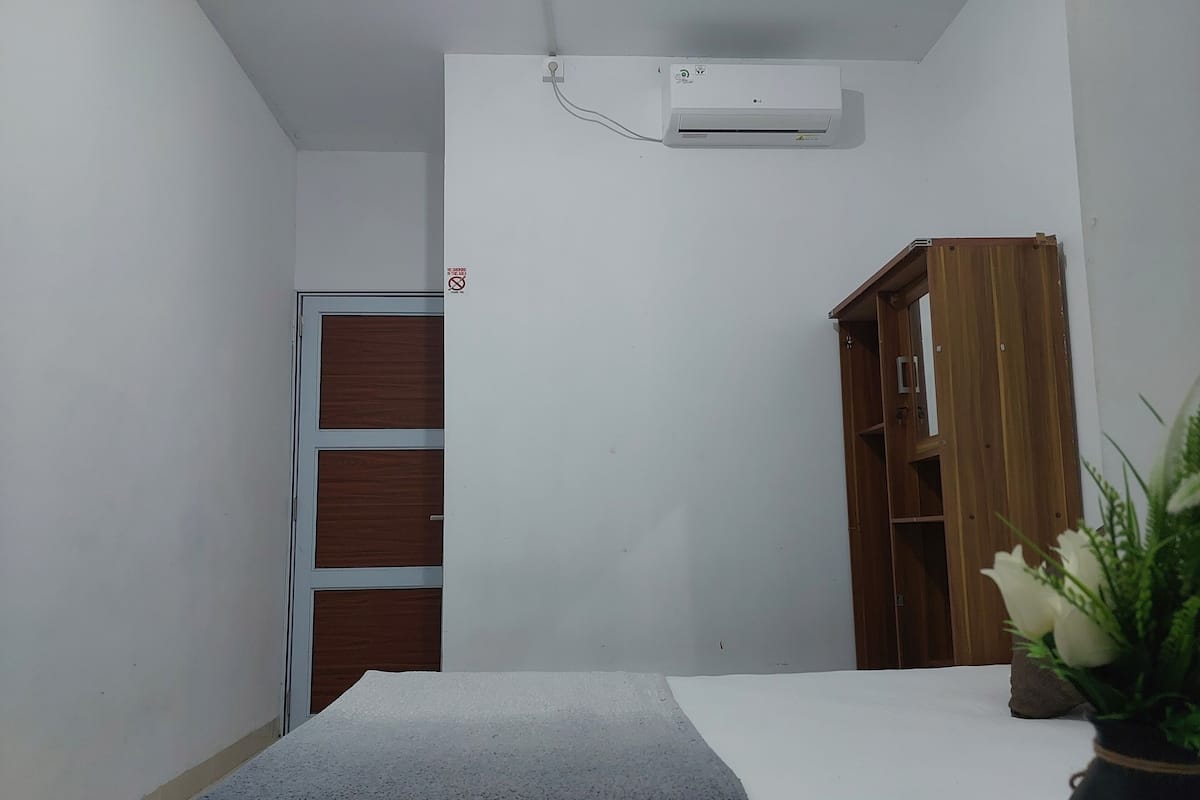 Ein kleiner, sauberer Raum mit einem weißen Bett, einem Holzschrank und einer weißen Klimaanlage an der Wand.