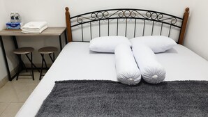 Ein ordentlich gemachtes Bett mit grauer Decke und Kissen, ein Holz-Kopfteil, ein kleiner Tisch mit zwei Hocker und eine Wasserflasche.