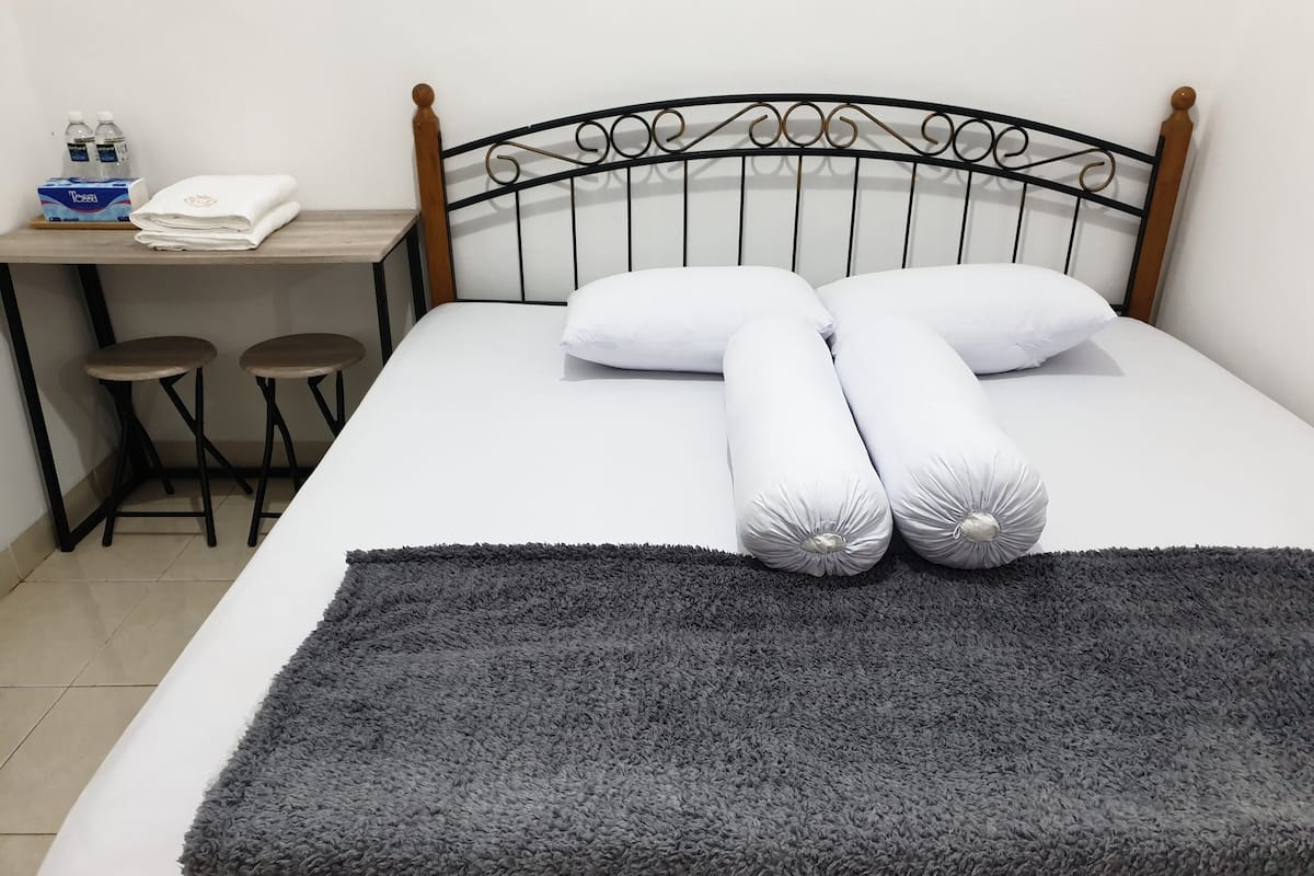 Ein ordentlich gemachtes Bett mit grauer Decke und Kissen, ein Holz-Kopfteil, ein kleiner Tisch mit zwei Hocker und eine Wasserflasche.