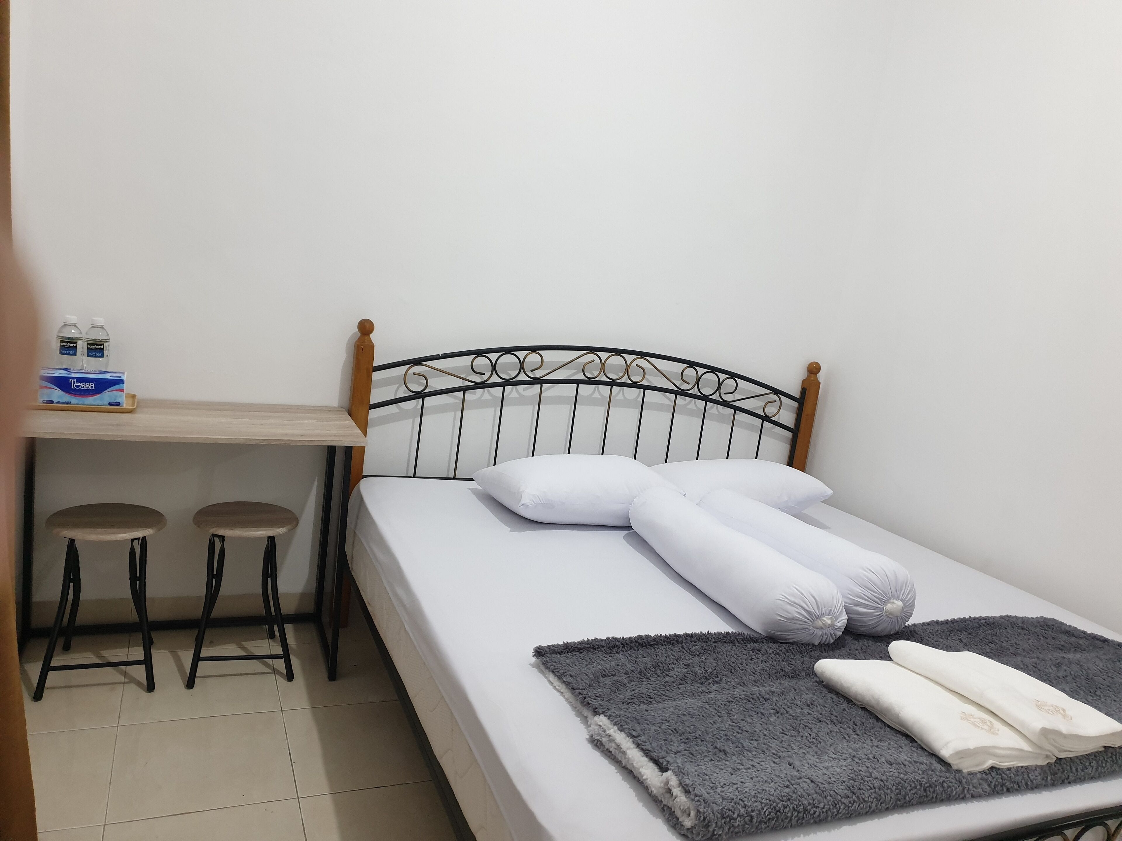 A small, neatly arranged room with a bed, a desk with two stools, and a shelf with bottled water.