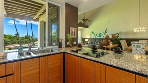 A modern kitchen with wooden cabinets, a granite countertop, and a large window overlooking a tropical landscape. - Palms At Wailea 2104 2 Bedroom Condo (Kihei)