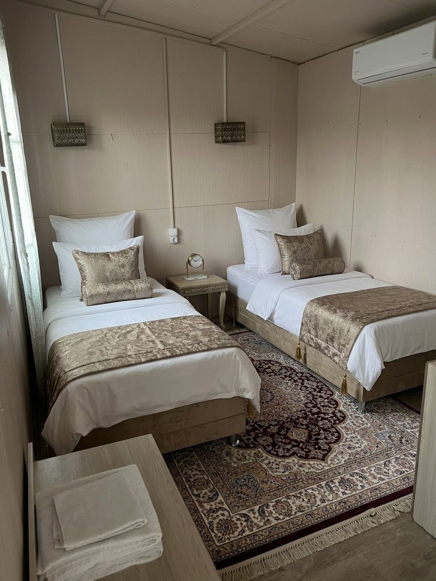 A hotel room with two beds, a large patterned rug, a bedside table with a lamp, and an air conditioning unit.