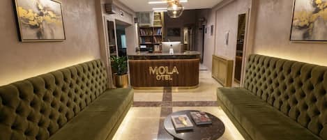 A lobby with a reception desk labeled 'MOLA HOTEL,' two green upholstered sofas, a marble coffee table, and a modern chandelier.