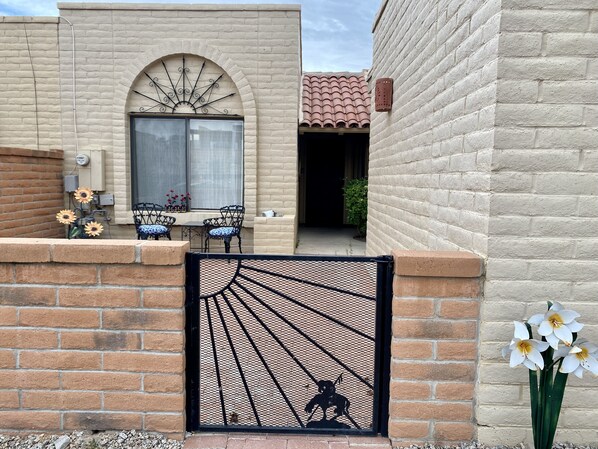 A brick building with a large arched window, a metal gate with a dog silhouette, and a small patio area with chairs and sunflowers. - Green Valley Casita - View ! (Green Valley)