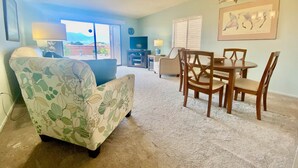 A living room with a floral armchair, a dining table with chairs, a chandelier, and a large window with a view. - Green Valley Casita - View ! (Green Valley)