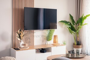A modern living room with a flat-screen TV mounted on a wooden wall, a wooden coffee table, a white console table with decorative items, and a large green plant. - Dieu Nérée & its sea view (Sète)