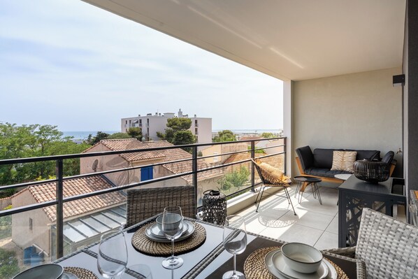 A balcony with a dining table set for two, a wicker chair, and a sofa with cushions. - Dieu Nérée & its sea view (Sète)