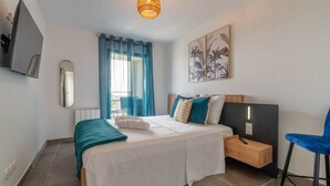 A bedroom with a bed, a bedside table, a chair, a TV, a mirror, and a window with curtains. - Dieu Nérée & its sea view (Sète)