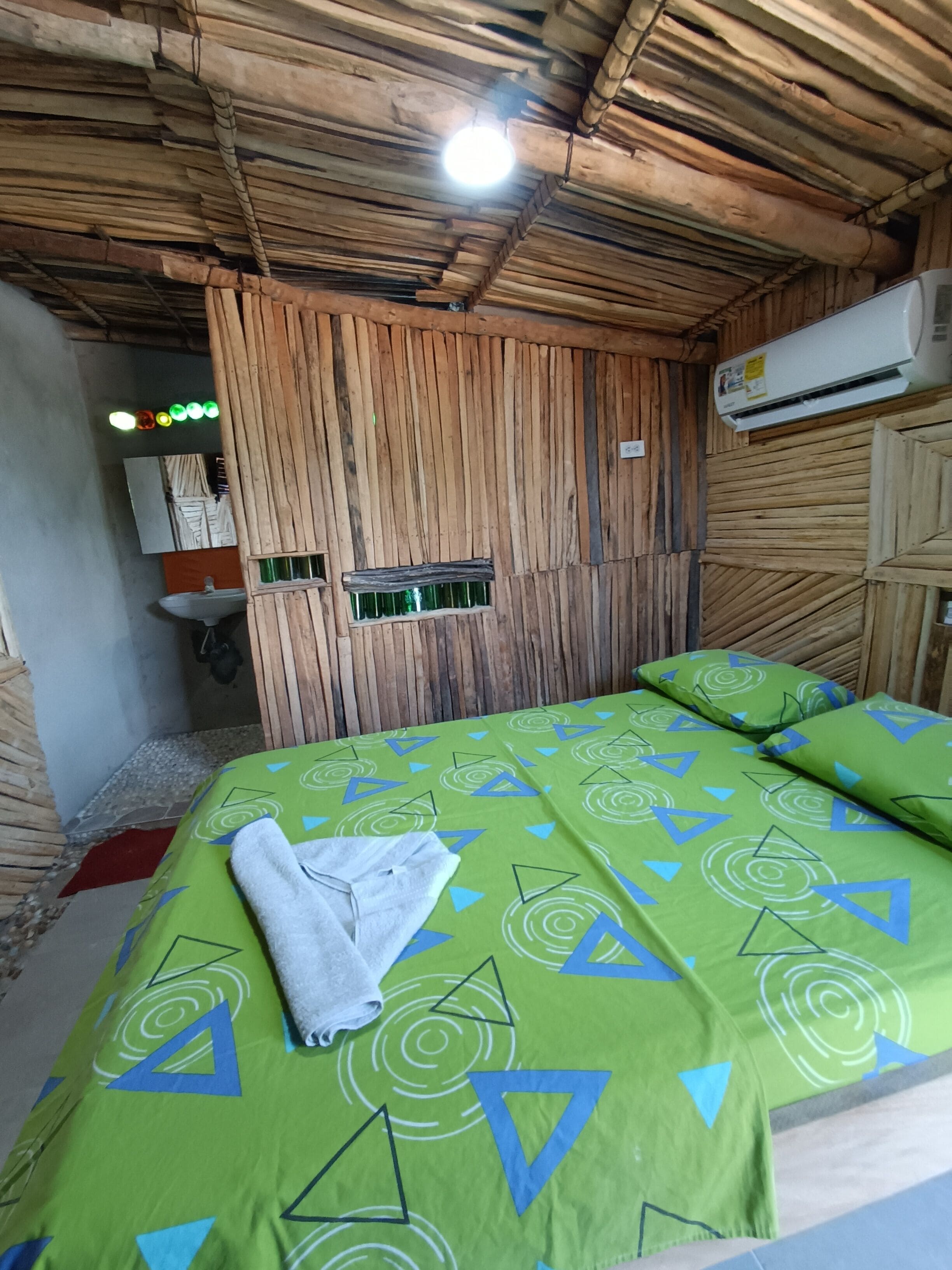 A room with a wooden ceiling, a bed with green patterned bedding, a white towel on the bed, a small window, and an air conditioning unit.