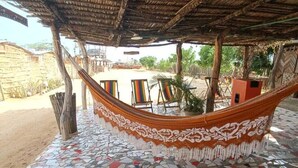 A hammock under a thatched roof, colorful mosaic flooring, and a sandy outdoor area with a building in the background. - Del mar vendra (Manaure)