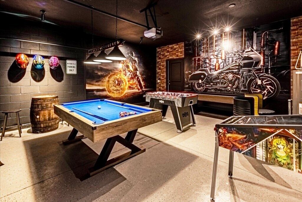 A billiards table, a foosball table, and a motorcycle-themed wall art in a room with a brick wall and a ceiling projector.