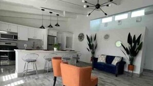 Eine moderne Küche mit weißen Schränken und einer zentralen Kochinsel, ein Essbereich mit orangefarbenen Stühlen und ein Wohnbereich mit einer Couch und Pflanzen.