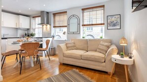 A modern living room with a beige sofa, a dining area with wicker chairs, and a kitchen in the background.