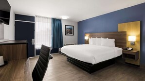 Habitación de hotel con una cama grande, un escritorio con silla, una mesita de noche con lámpara y una ventana con cortinas.