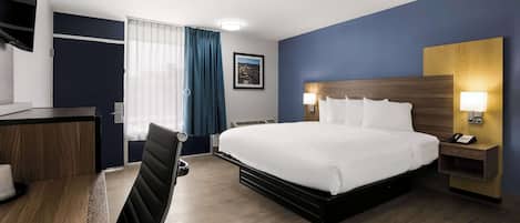 Ein Hotelzimmer mit einem großen Bett, einem Schreibtisch mit Stuhl, einem Nachttisch mit Lampe und einem Fenster mit Vorhängen.