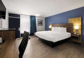 A hotel room with a large bed, a desk with a chair, a bedside table with a lamp, and a window with curtains.