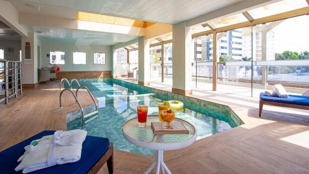 A modern indoor swimming pool with clear glass walls, a sunlit lounge area, and a view of the cityscape.