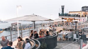 A waterfront restaurant with outdoor seating, a view of the water, and a modern building in the background. - Trendy Getaway | Hot Tub | 20 mins to Disney (Long Beach)