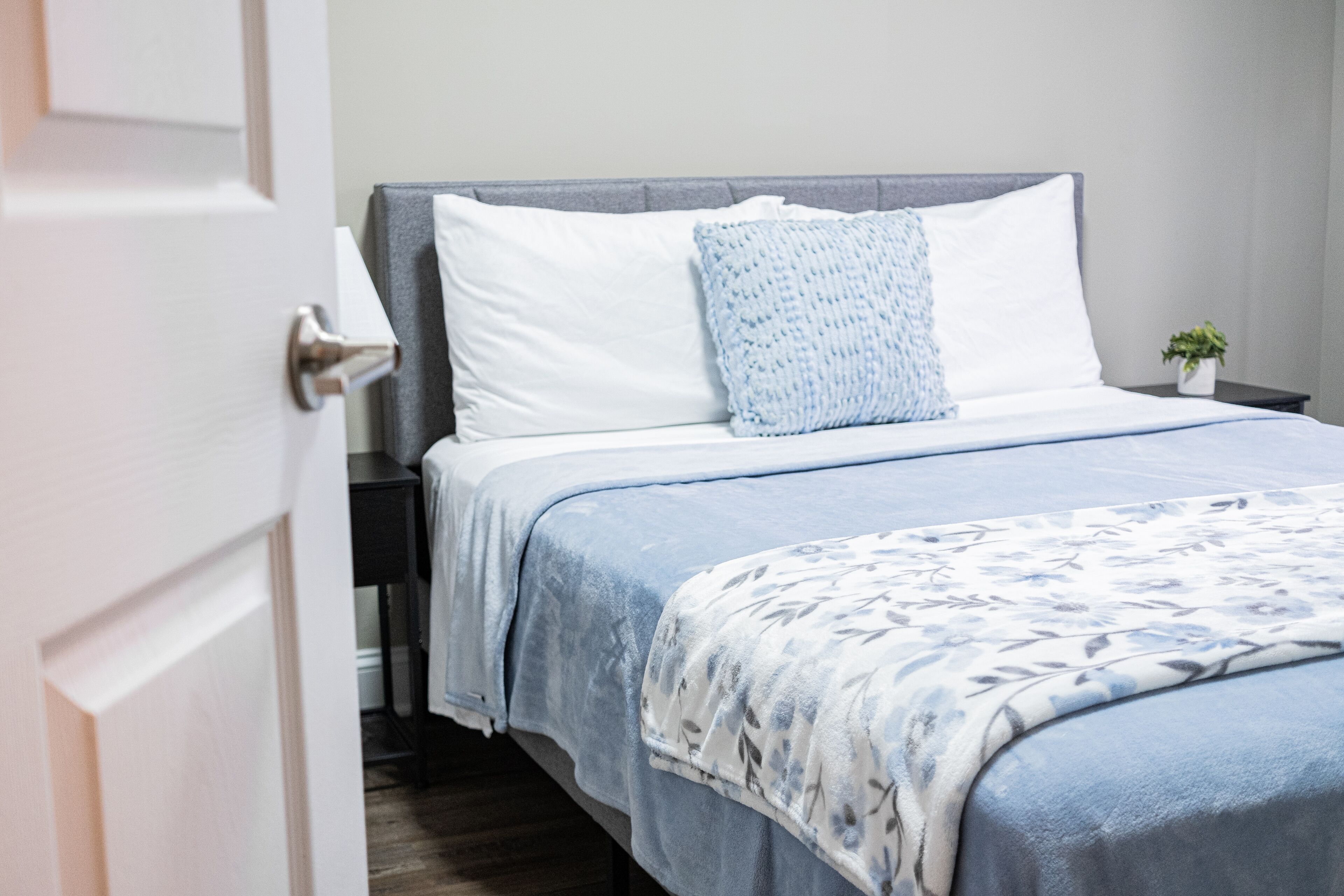 A neatly made bed with a blue and white floral bedspread, a white pillow, and a blue knitted throw.