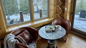 A cozy cabin interior with a wooden ceiling, a round table with a puzzle, a leather chair, and a view of a snowy forest through large windows. - Ranch Creek Log Home (Fraser)
