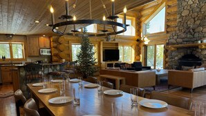 A spacious dining area with a long wooden table set for a meal, a stone fireplace, and a cozy seating area with a television. - Ranch Creek Log Home (Fraser)