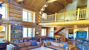 A spacious log cabin interior with a central seating area, a dining table, and a staircase leading to an upper level. - Ranch Creek Log Home (Fraser)