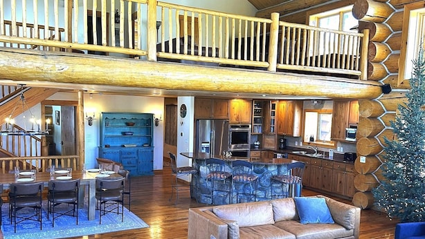 A spacious living area with a high ceiling, wooden beams, a dining area, and a staircase leading to an upper level. - Ranch Creek Log Home (Fraser)