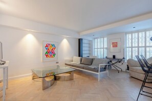 A modern living room with a glass coffee table, a sofa, and a chair. There are artworks on the walls and a window with blinds.
