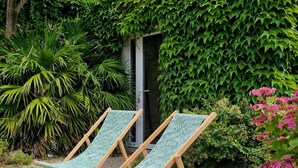 A garden with two wooden lounge chairs, a gravel path, and lush greenery including a palm tree and ivy-covered wall.