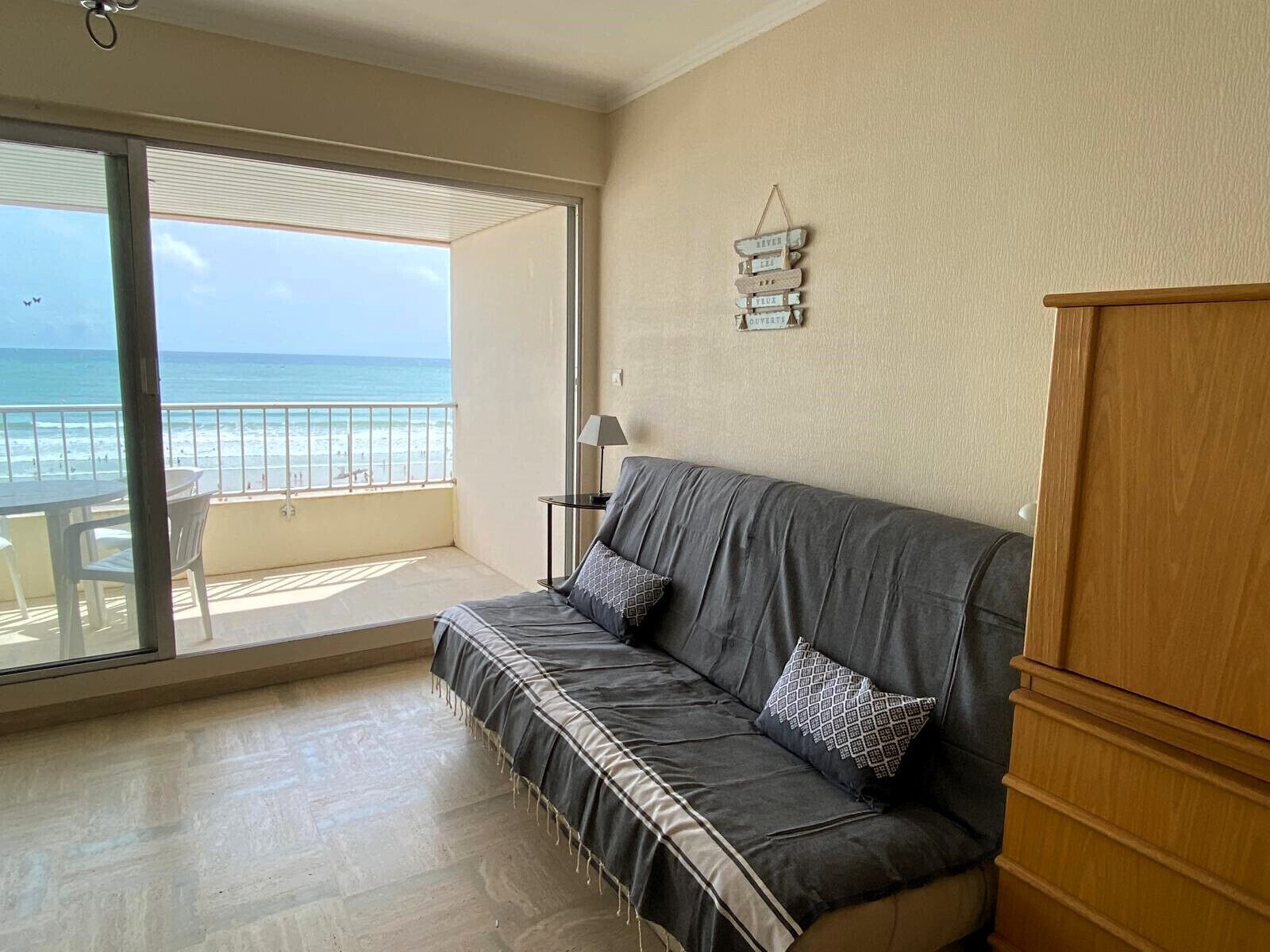 Un balcón con vista al mar, un sofá gris con cojines estampados y un armario de madera.