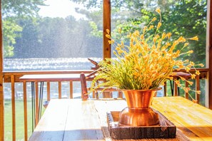 A wooden table with a copper vase containing yellow flowers, set on a wooden deck with a view of a lake and trees. - Welcome to Dells Escape: A Historic Getaway on Dells Mill Pond (Augusta)