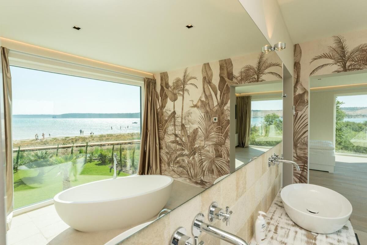 A modern bathroom with a large oval sink, a mirror, and a view of the beach.