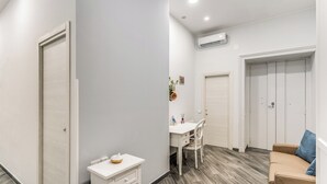 Una habitación moderna y luminosa con piso de madera, un escritorio blanco con una silla, un sofá color beige y un armario blanco.