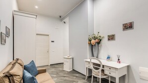 Una habitación moderna con un sofá color beige, un escritorio blanco y una silla. Se observa un mueble pequeño y una puerta al fondo.