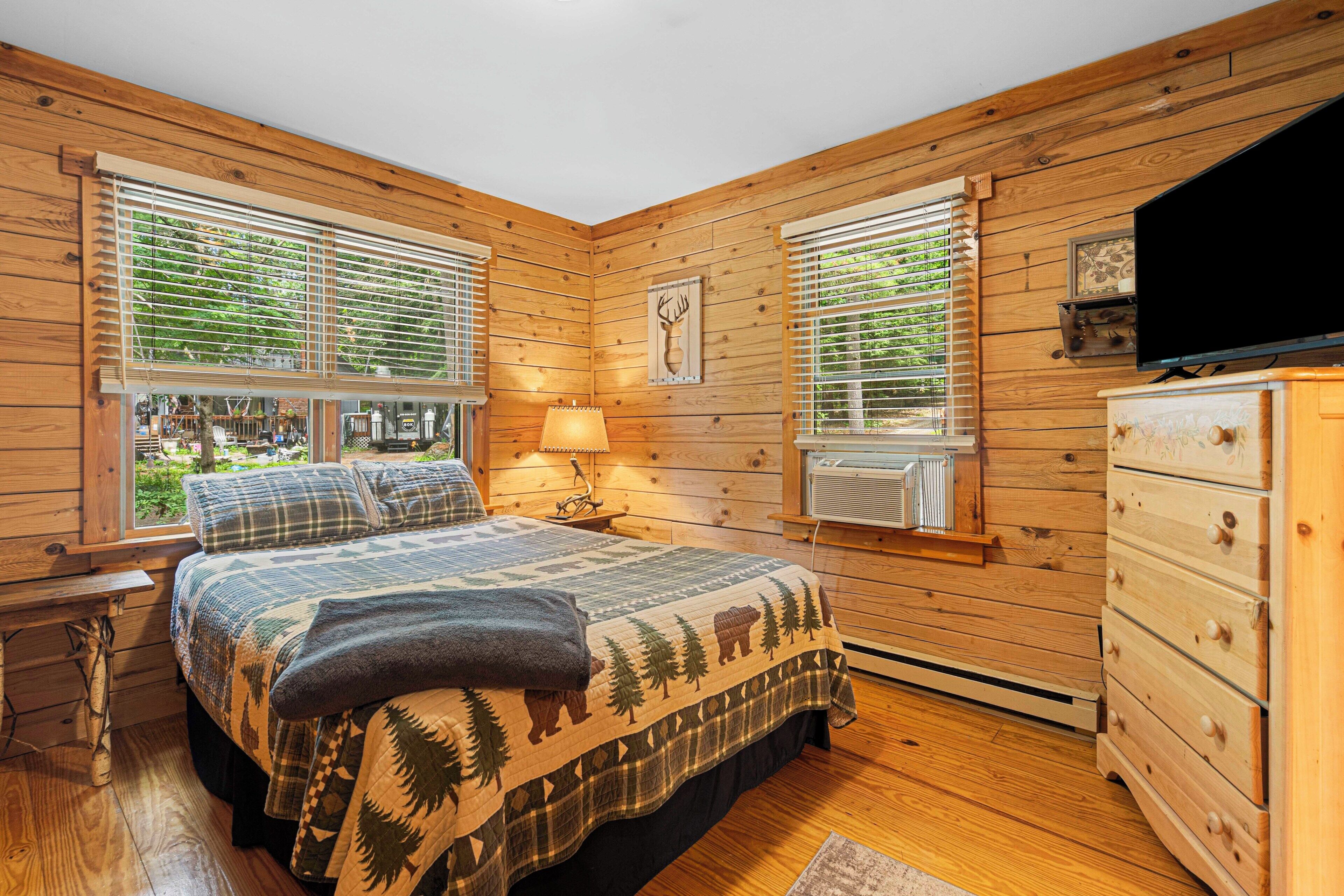 Une chambre dans une cabane en bois, avec un lit, une commode et deux fenêtres équipées de stores.