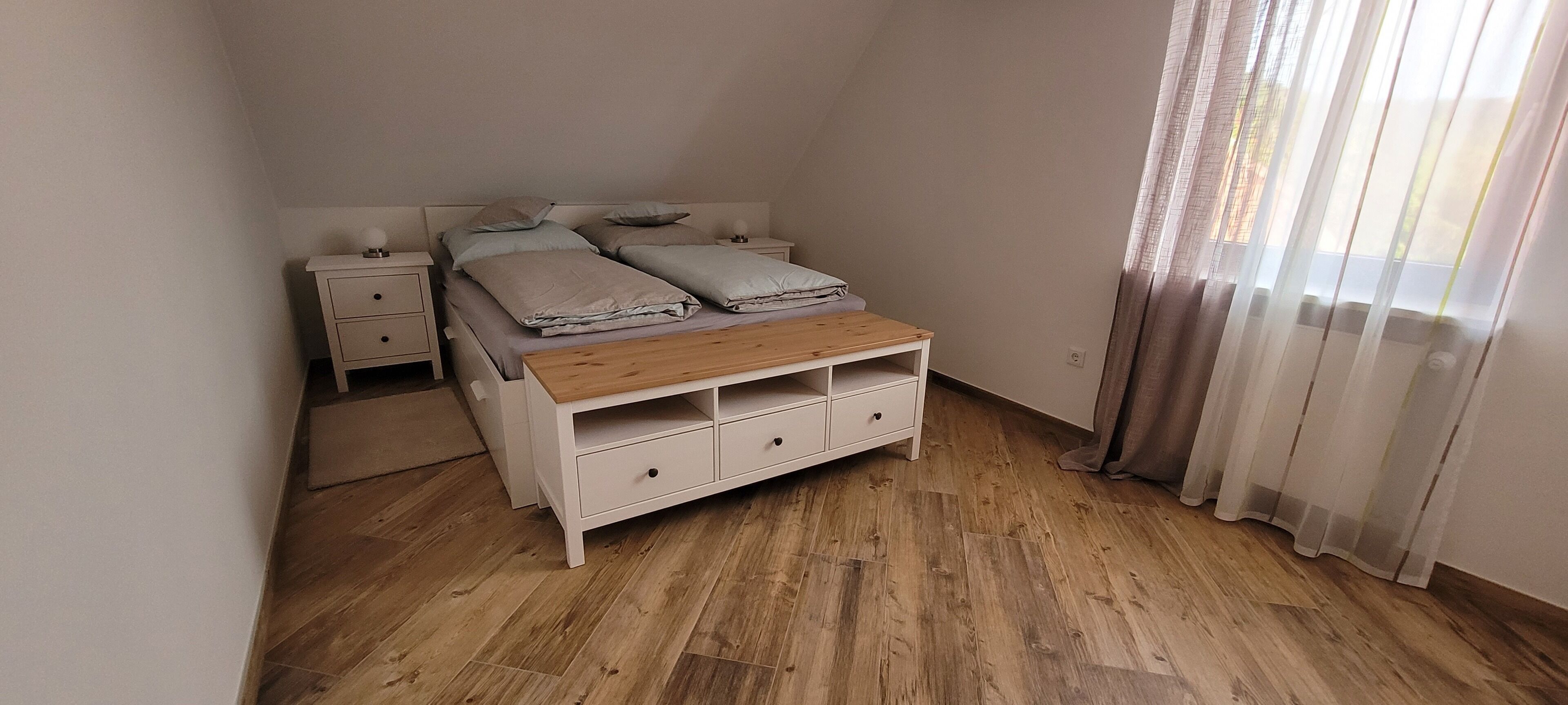 Ein Schlafzimmer mit einem hölzernen Bett, zwei Nachttischen, einem Schminktisch und einem Holzschrank, sowie durchsichtigen Vorhängen.