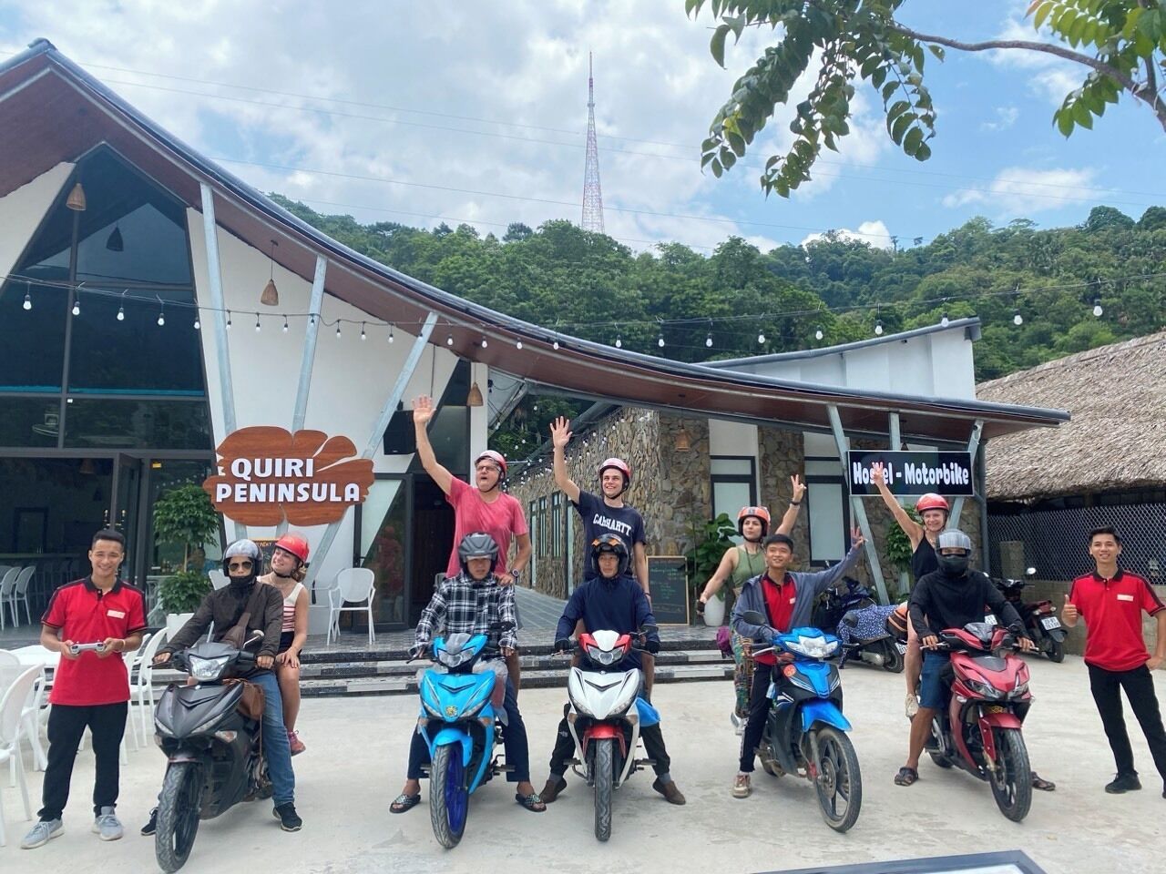 A group of people on motorcycles in front of a building with a sign reading "Quiri Peninsula Hotel - Motorbike".
