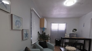 Un salón con un sillón azul claro, una mesa de centro de vidrio y una mesa de comedor con sillas. En la pared hay cuadros enmarcados y una planta en el alféizar de la ventana.