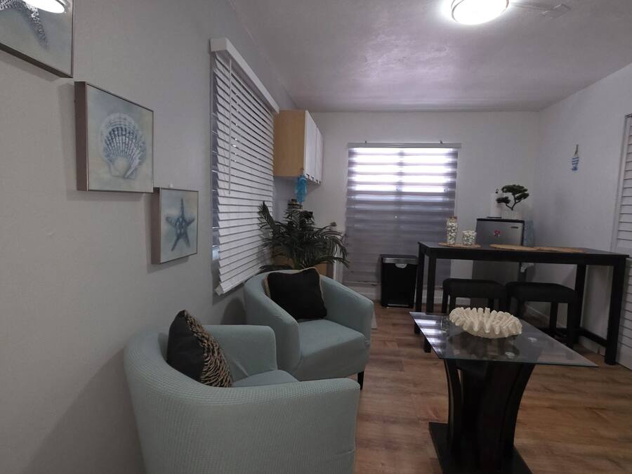 Un salón con un sillón azul claro, una mesa de centro de vidrio y una mesa de comedor con sillas. En la pared hay cuadros enmarcados y una planta en el alféizar de la ventana.