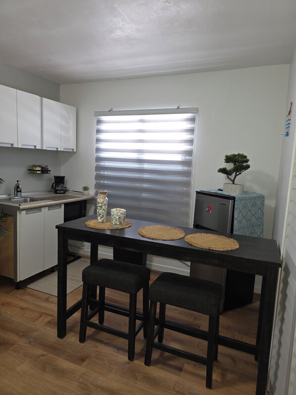 Una cocina compacta con armarios blancos, una mesa de comedor negra con dos sillas, una cafetera y una pequeña planta en maceta.