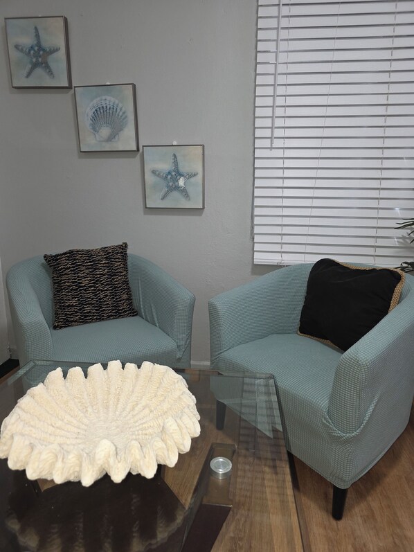 Un salon avec deux fauteuils turquoise, une table basse en verre et trois œuvres d’art encadrées accrochées au mur.